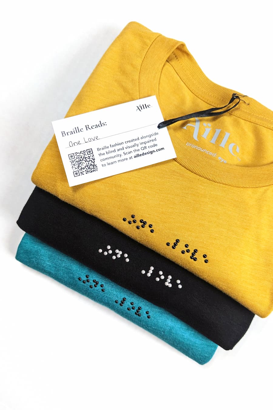 Close up of braille clothing: yellow shirt with black braille, black shirt with white braille, teal shirt with black braille