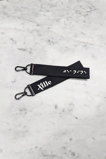 Black keychain with white braille on one side and a white screen printed Aille Design logo on the other side.