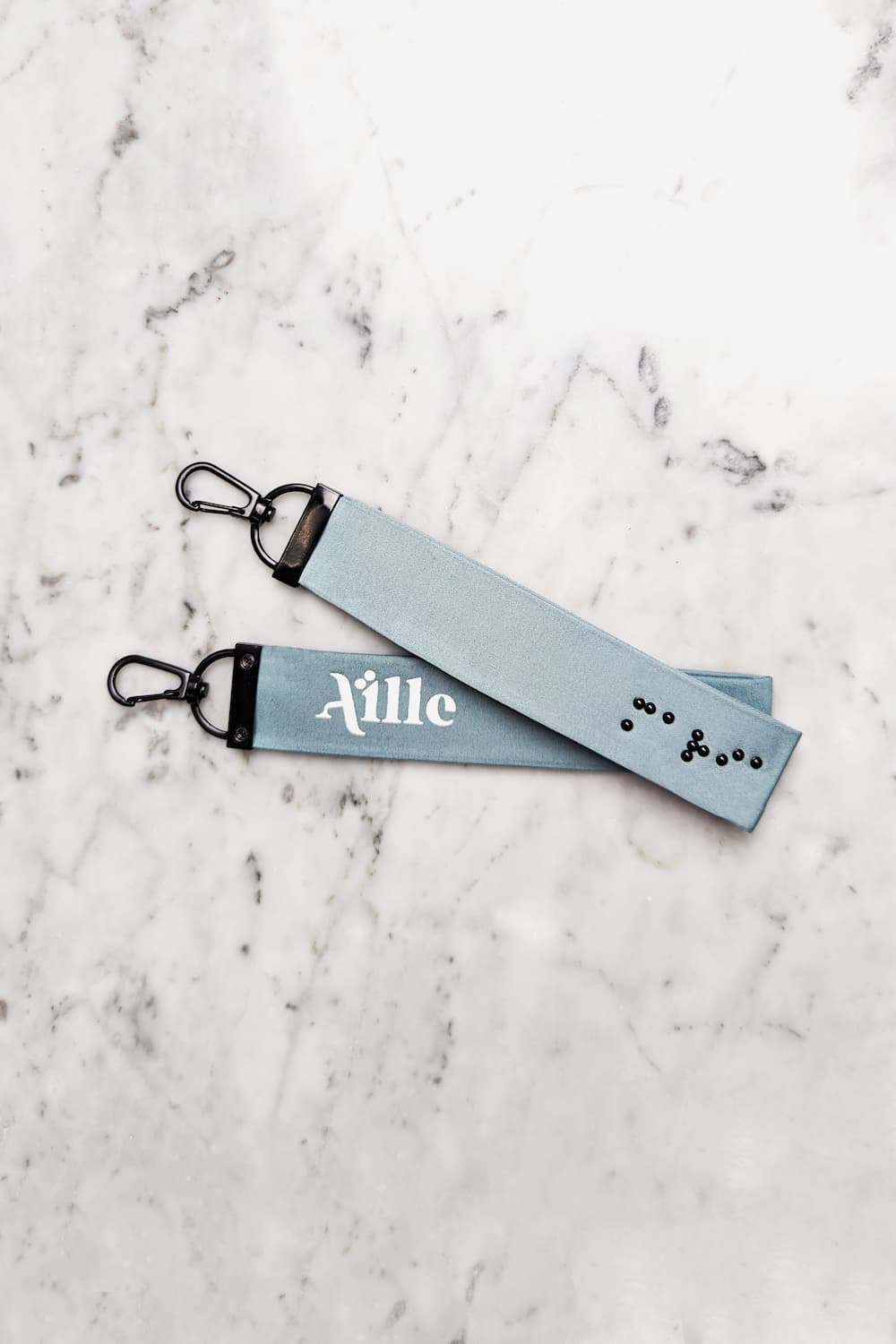 Light blue/grey keychain with black braille on one side and a white screen printed Aille Design logo on the other side.