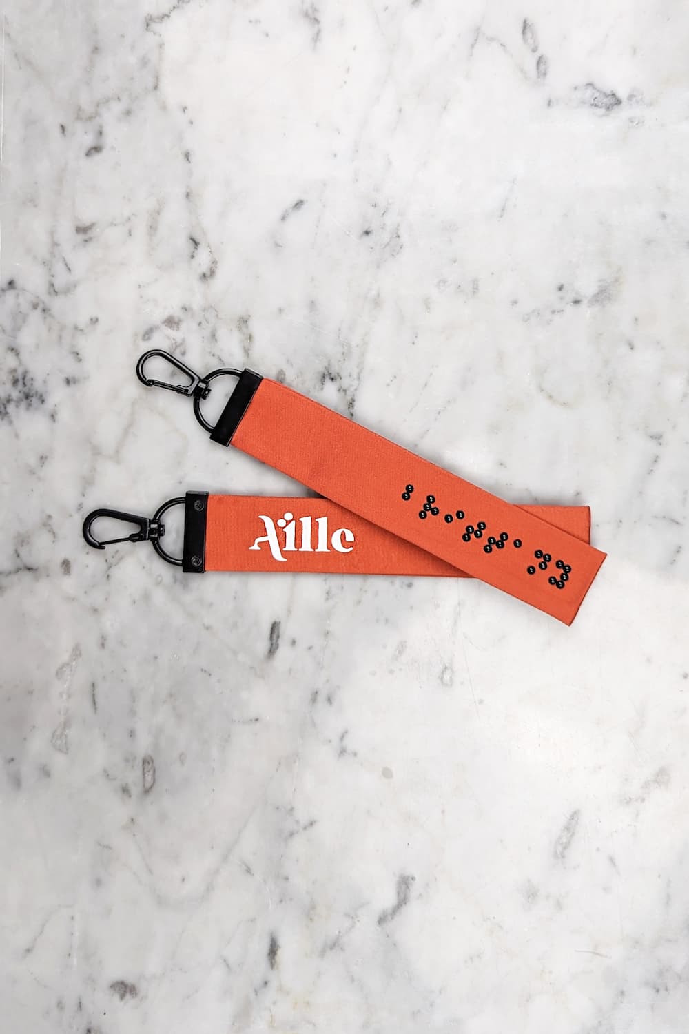 Orange keychain with black braille on one side and a white screen printed Aille Design logo on the other side.