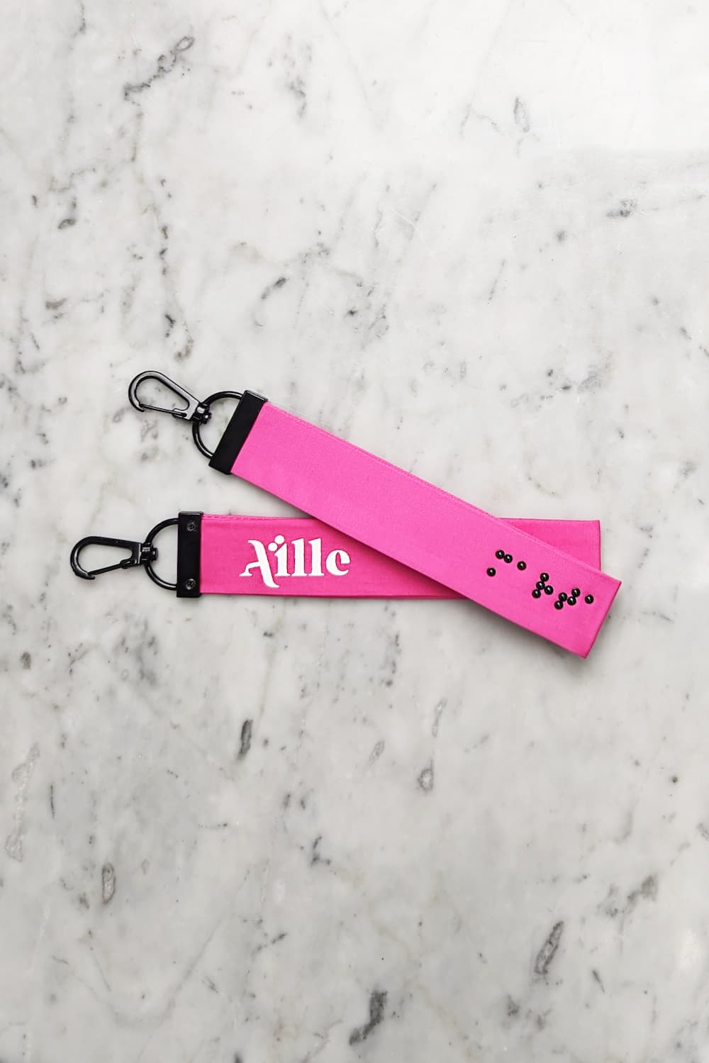 Hot pink keychain with black braille on one side and a white screen printed Aille Design logo on the other side.