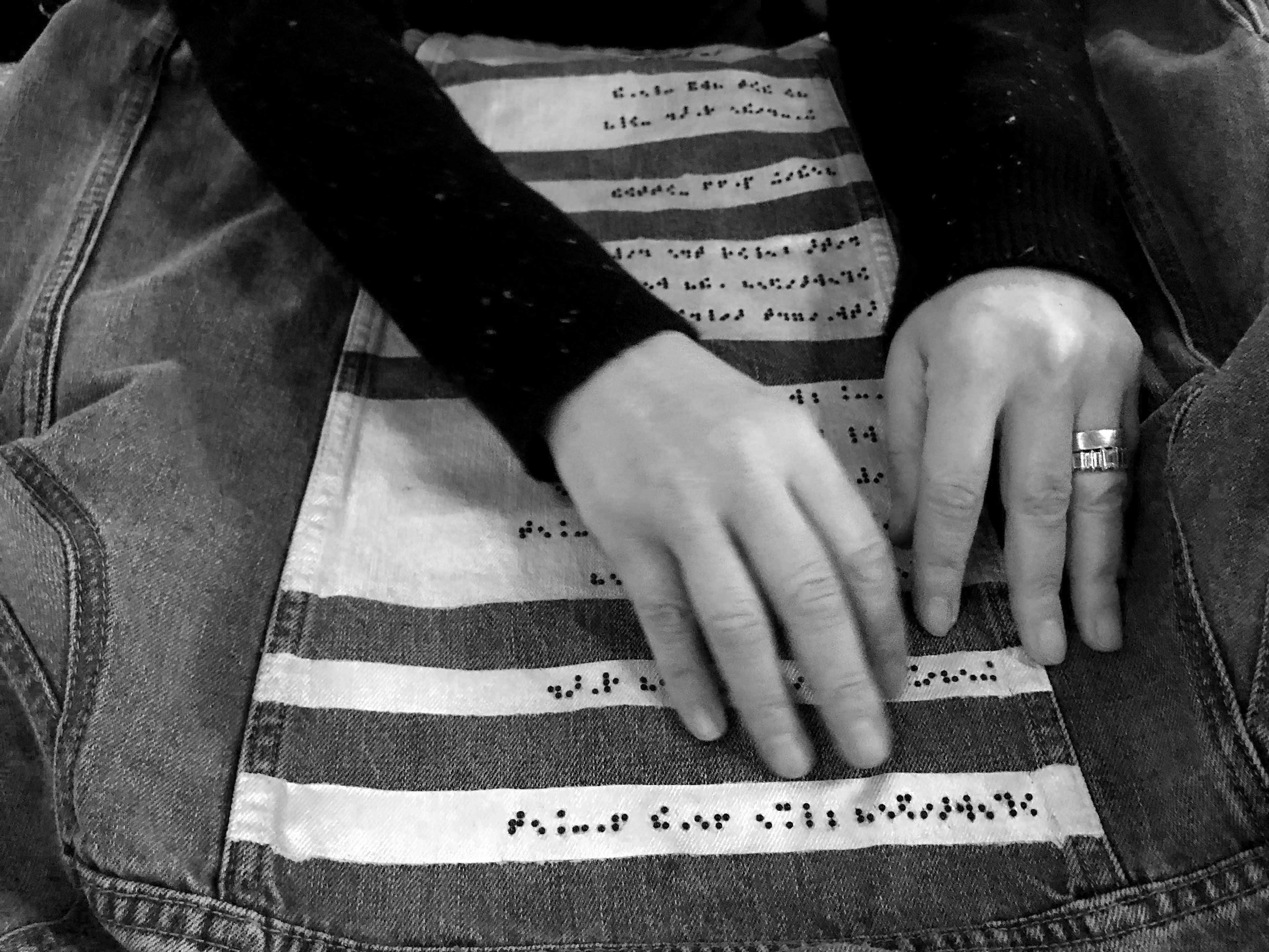 Black and white close up photo of a woman's hands reading the black braille beadwork on our denim jacket prototype.