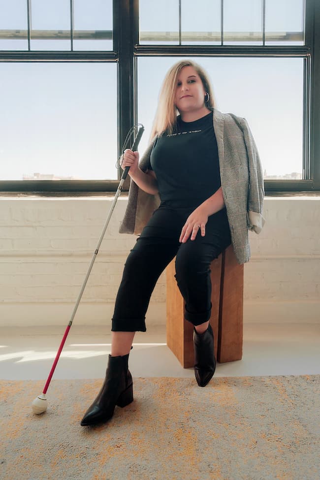 Visually impaired white woman holding her white cane. She is wearing a black braille t-shirt and blazer.