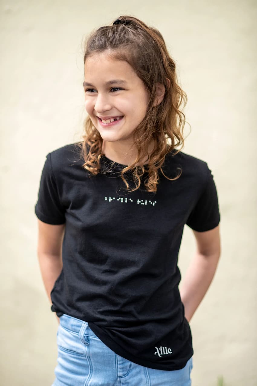 A young girl is wearing a braille t-shirt and is smiling and looking away from the camera.