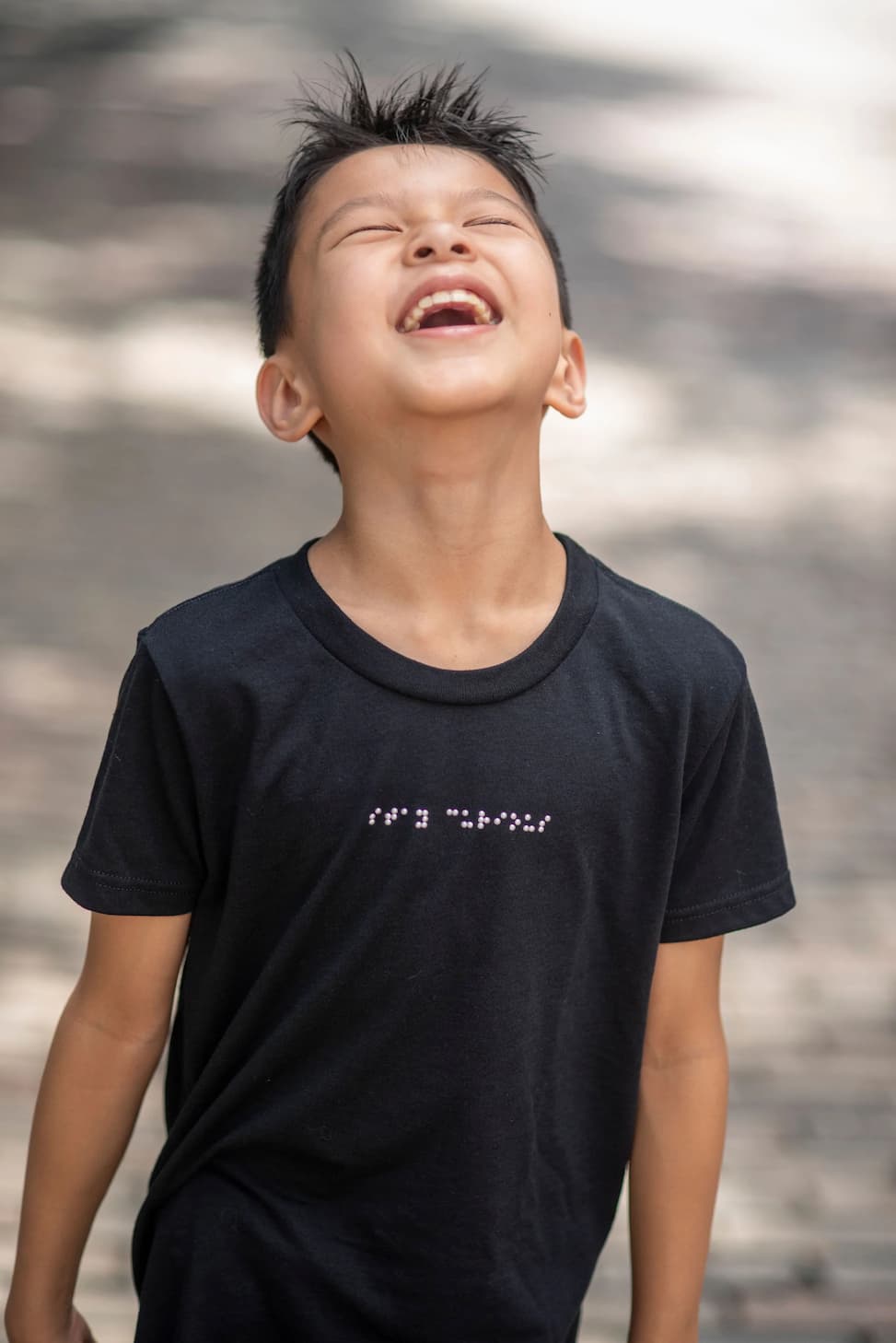 A young boy is wearing a braille t-shirt, looking up, and has the biggest smile on his face.