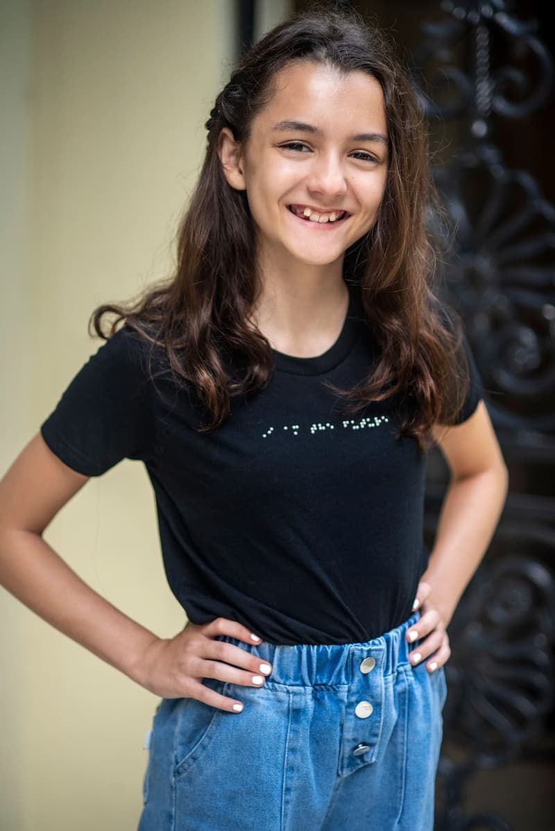 A young girl is wearing a braille t-shirt, has her hands on her hips, and has a beautiful smile.