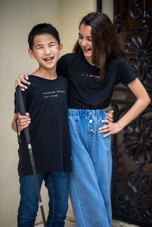 A young boy with a white cane standing next to his sister. Both kids are wearing braille t-shirts and laughing.