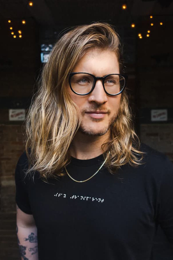 Young man with long hair, glasses, tattoos and piercings wearing custom braille clothing
