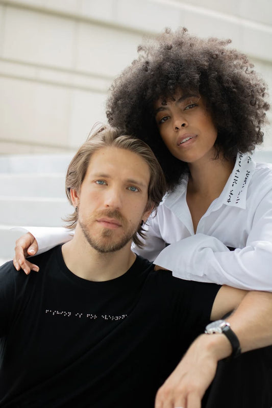 Close up of white man and Black Latina woman sitting together. Man is wearing black braille t-shirt. Woman is wearing white collared shirt with braille.