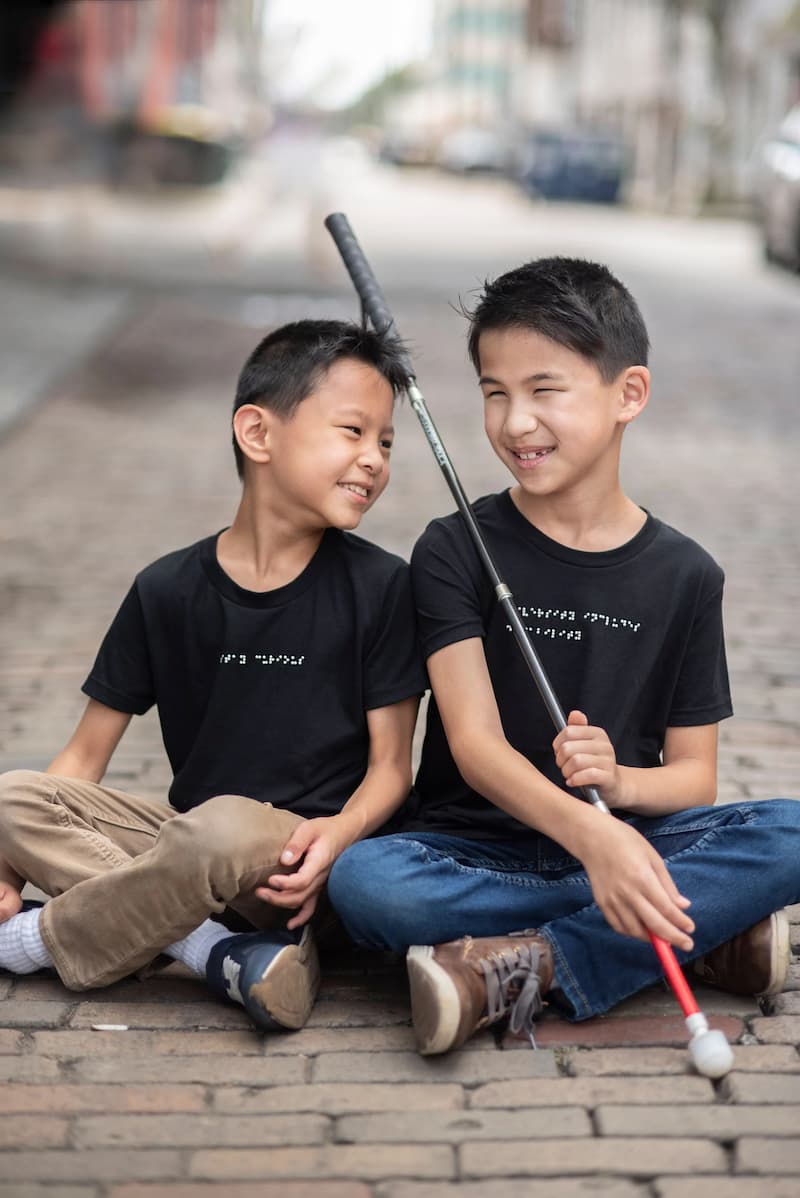 Two young boys are sitting on a cobble stone road. They are both wearing braille t-shirts, and looking at each other smiling. One boy has a white cane.