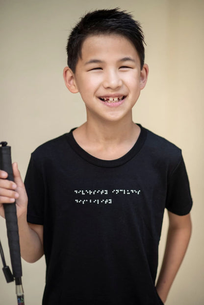 A young boy with a white cane is wearing a braille t-shirt and smiling at the camera.