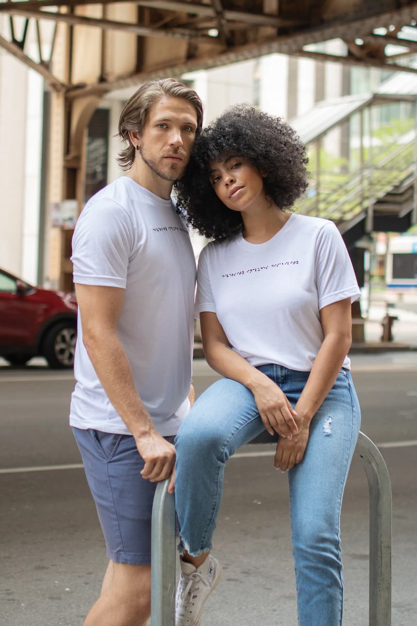White man and Black Latina woman standing together wearing white braille t-shirts