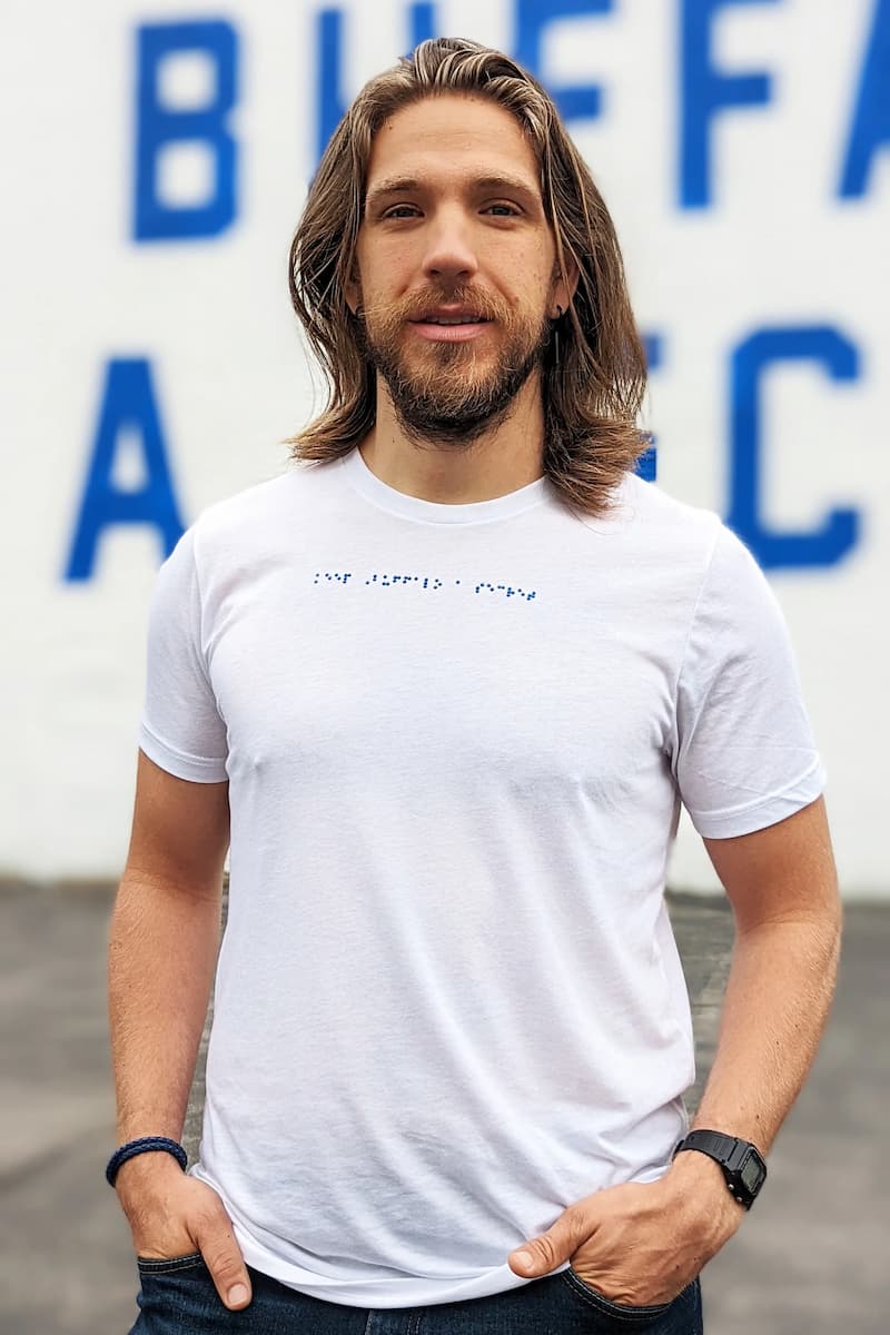 Jake is standing in front of a mural with his hands in his pockets. He is wearing a white t-shirt with blue braille