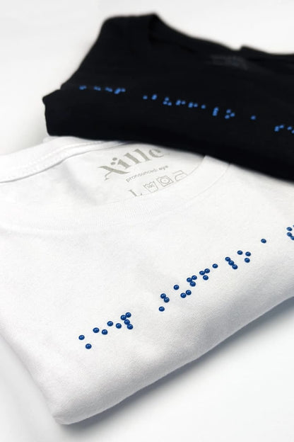 One black and one white braille t-shirt folded on a white table. Both t-shirts have blue braille.
