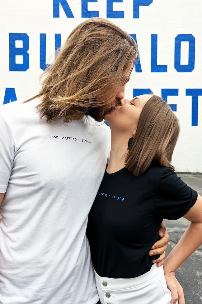 Jake and Alexa kissing in front of Keep Buffalo A Secret mural while wearing their braille t-shirts.