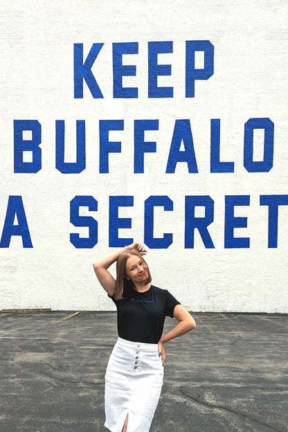 Alexa is standing in front of a mural that reads "Keep Buffalo A Secret". She is wearing a black t-shirt with blue braille and a white denim pencil skirt.