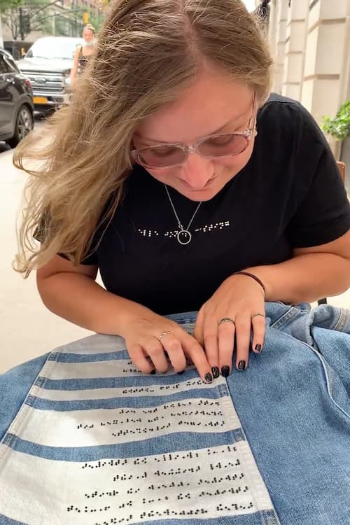 White visually impaired woman wearing black braille t-shirt while reading braille on braille denim jacket
