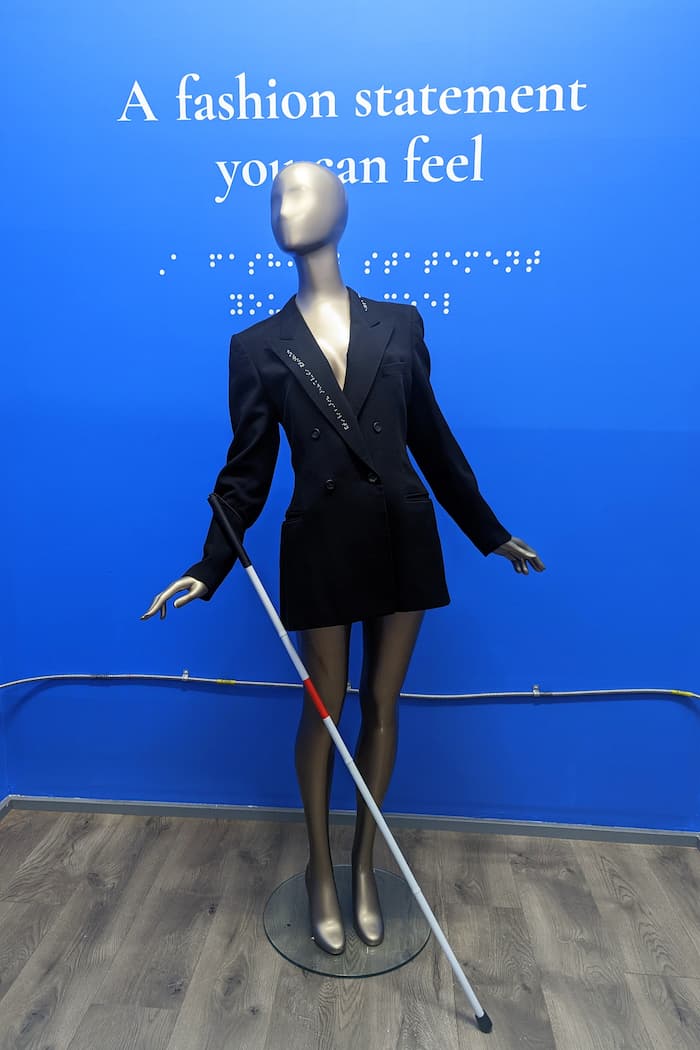Mannequin with white cane wearing black blazer with chrome braille